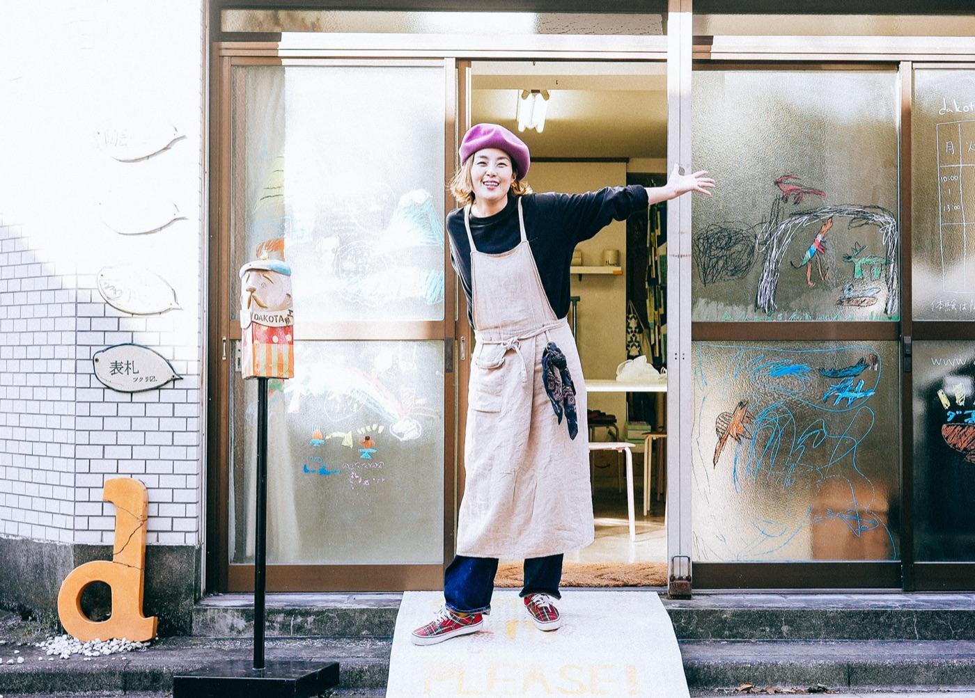 ベレー帽の講師が店先で腕を広げて出迎える様子。ガラス戸には子供が描いた絵が残る
